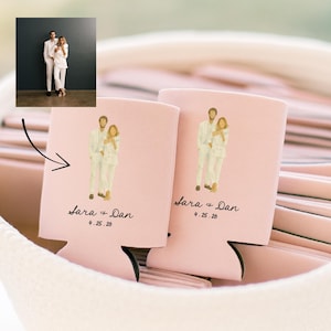 May include: Two pink can coolers with a wedding illustration. Each cooler features a couple in white suits, the names "Sara + Dan", and the date "4.25.26" in black script. The coolers are nestled in a soft, pink fabric.