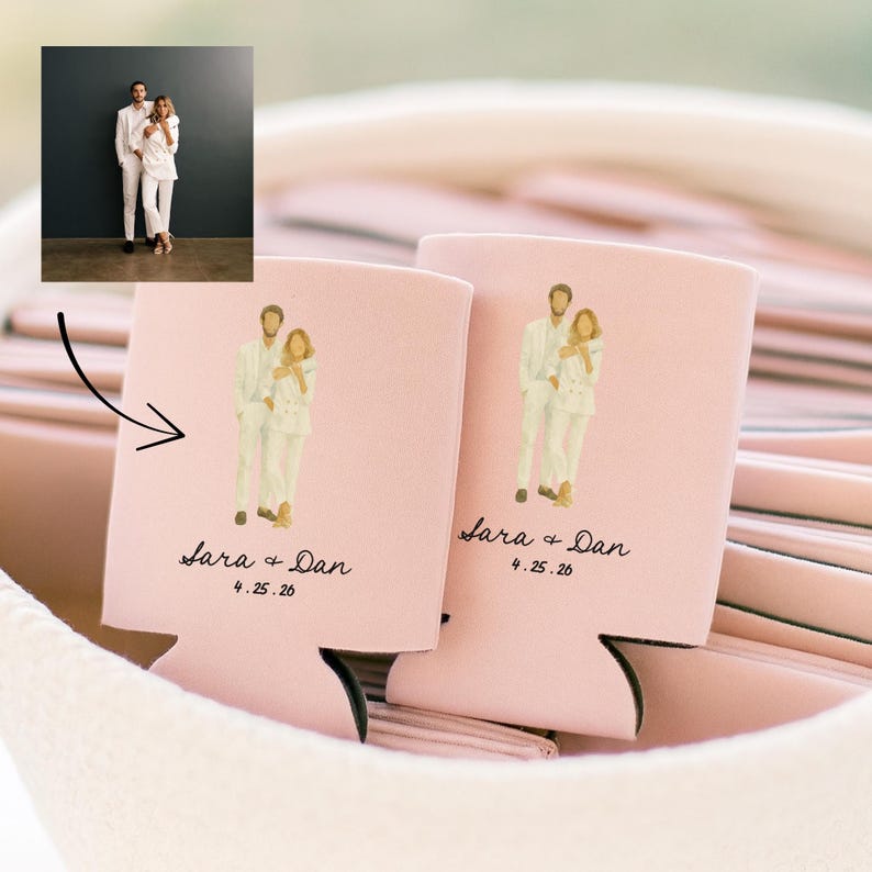 May include: Two pink can coolers with a wedding illustration. Each cooler features a couple in white suits, the names "Sara + Dan", and the date "4.25.26" in black script. The coolers are nestled in a soft, pink fabric.