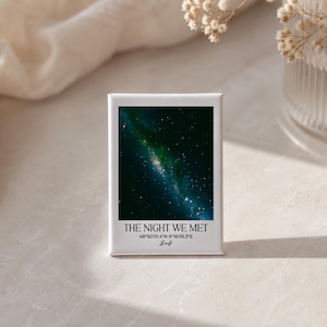 May include: A rectangular white magnet featuring a dark night sky image with stars and a green nebula. The text reads THE NIGHT WE MET, followed by the coordinates 48°50'01.4"N 9°16'05.3"E and the cursive initials L&P printed in black ink.