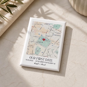 Könnte beinhalten: Eine weiße Keramikfliese mit einer Stadtkarte und einem roten Herz, das einen Ort markiert. Der Text "OUR FIRST DATE" steht unter der Karte, zusammen mit dem Datum 15.05.2022, den Koordinaten und den Namen "Roah & Paul".