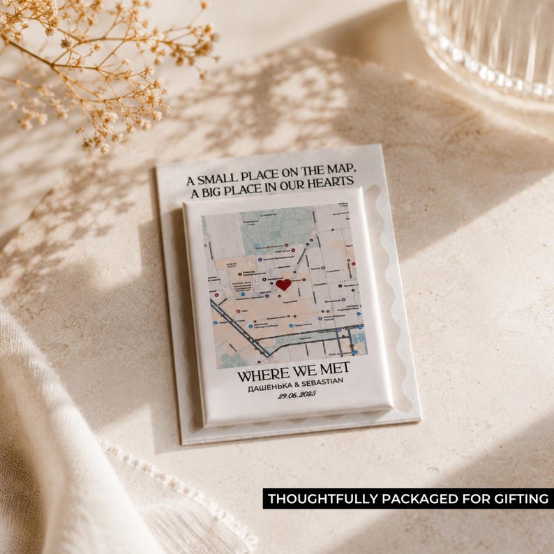 May include: A white rectangular magnet featuring a map design with a red heart marking a location. The text "WHERE WE MET" and names are below the map. The magnet is on a white card with the text "A SMALL PLACE ON THE MAP, A BIG PLACE IN OUR HEARTS".