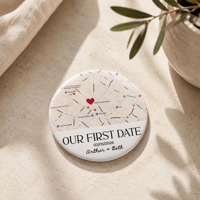 May include: A circular white pinback button featuring a printed street map with a red heart icon marking a specific location. Below the map, the text reads OUR FIRST DATE, 02/05/2026, and the names Arthur & Beth in a black serif and script font.