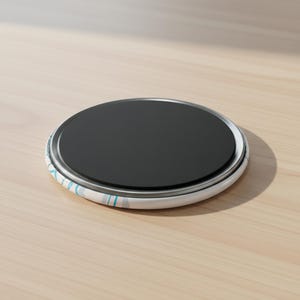 May include: A round magnet with a black center and a white and blue patterned border. The magnet is sitting on a light brown wooden surface.