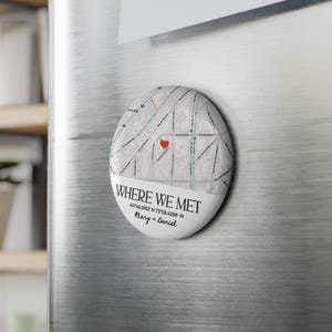 May include: An oval magnet with a map design, featuring a red heart marking a location. The text "WHERE WE MET" is displayed, along with coordinates and names. The magnet is attached to a metallic surface.