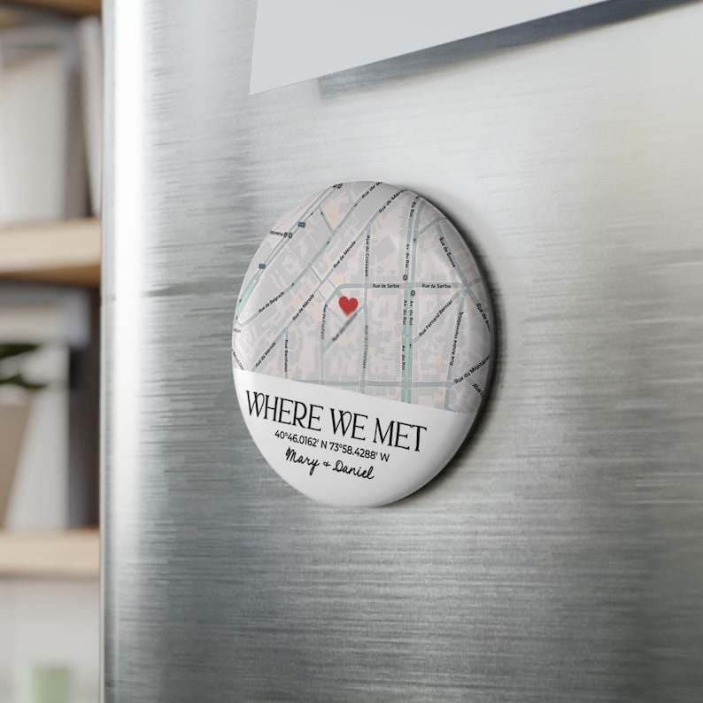 May include: An oval magnet with a map design, featuring a red heart marking a location. The text "WHERE WE MET" is displayed, along with coordinates and names. The magnet is attached to a metallic surface.