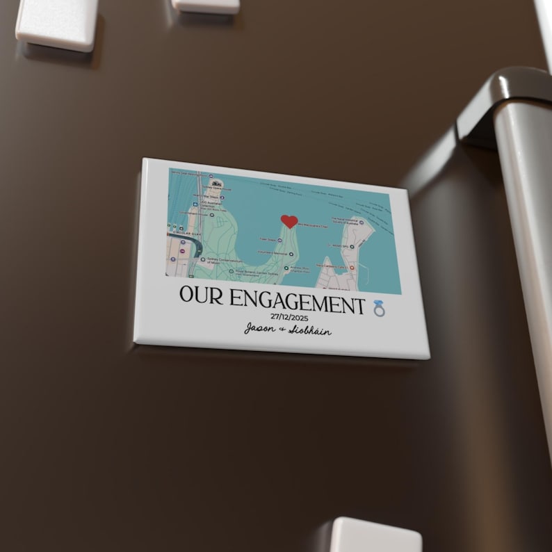 May include: A white rectangular magnet with the words "OUR ENGAGEMENT" and the date 27/12/2025. The magnet features a map with a red heart marking a location. The names "Jason + Siobhain" are below the date.