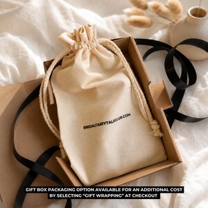 May include: A beige drawstring bag with the text "BRIDALFAIRYTALECLUB.COM" inside a cardboard box, with a black ribbon and decorative elements. The bag is made of a soft, natural fabric.