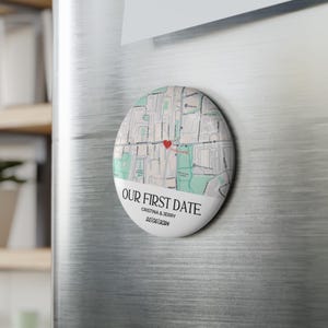 May include: A round magnet with a map design and the text "OUR FIRST DATE". The map features a red heart marking a location. Below the map is the text "CRISTINA & JERRY 25/06/2024". The magnet is attached to a silver refrigerator.