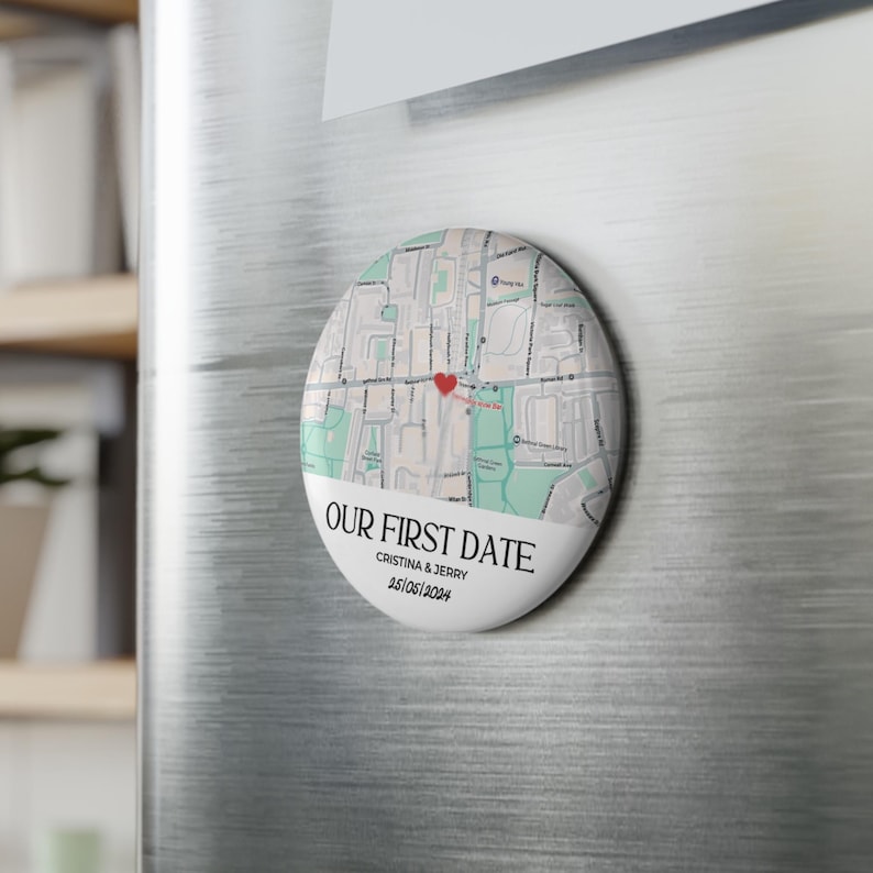 May include: A round magnet with a map design and the text "OUR FIRST DATE". The map features a red heart marking a location. Below the map is the text "CRISTINA & JERRY 25/06/2024". The magnet is attached to a silver refrigerator.