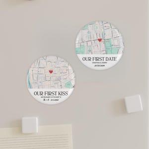 May include: Two round magnets with map designs. One says "OUR FIRST KISS" with coordinates and a date. The other says "OUR FIRST DATE" with names and a date. Both have a red heart marker.