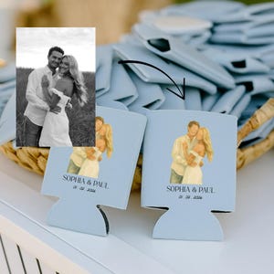 May include: Light blue can coolers featuring a watercolor illustration of a couple embracing. The text "SOPHIA & PAUL" and the date "01. 02. 2026" are printed below the illustration. A black and white photo of the couple is also visible.