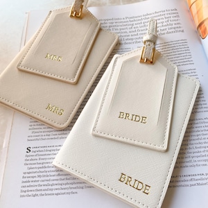 Peut inclure: &Eacute;tiquettes de bagages ivoire avec des lettres dor&eacute;es "MRS" et "BRIDE". Les &eacute;tiquettes ont une forme rectangulaire avec une petite &eacute;tiquette sur le dessus et une boucle dor&eacute;e. Les &eacute;tiquettes sont sur un magazine.