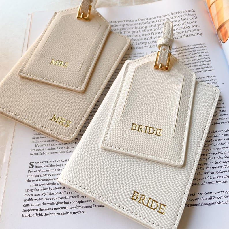 Peut inclure: &Eacute;tiquettes de bagages ivoire avec des lettres dor&eacute;es "MRS" et "BRIDE". Les &eacute;tiquettes ont une forme rectangulaire avec une petite &eacute;tiquette sur le dessus et une boucle dor&eacute;e. Les &eacute;tiquettes sont sur un magazine.