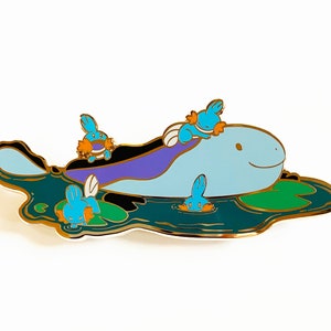 May include: A gold-coloured enamel pin featuring a blue and purple cartoon water Pokémon with three smaller blue and orange Pokémon swimming around it. The larger Pokémon is on a green lily pad with a gold outline.