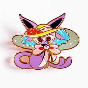 Puede incluir: Un pin de personaje de dibujos animados morado con ribete dorado, que lleva un sombrero amarillo con rosas rojas y un lazo rosa. El personaje tiene ojos grandes y está sobre un fondo blanco. El diseño es caprichoso y decorativo.