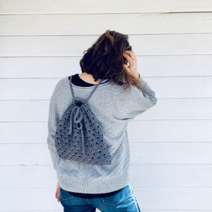 May include: A person wearing a gray sweater and blue jeans is wearing a gray crocheted drawstring backpack.