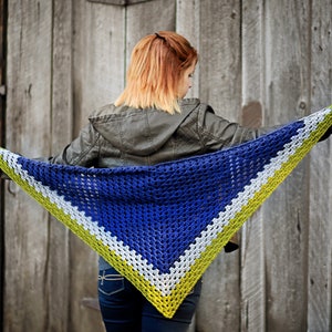 May include: A blue, white and green crocheted triangular shawl with a fringe. The shawl is being held by a person wearing a brown leather jacket and blue jeans.