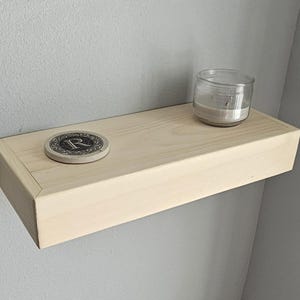 May include: A light-colored wooden floating shelf mounted on a gray wall. A coaster with an ornate "R" design and a small glass jar candle sit on the shelf. The shelf is a simple, rectangular design.
