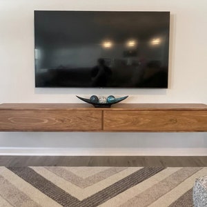 May include: A wall-mounted, light brown wooden entertainment center with two drawers, below a large black television. A decorative black bowl with blue and silver spheres sits on top. The unit is mounted on a white wall.