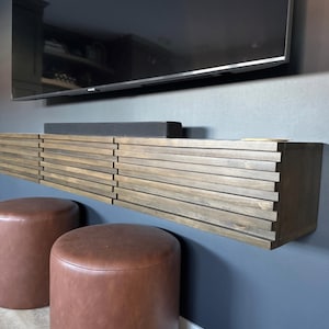 May include: A floating wooden media console with a slatted design is mounted on a dark blue wall. Two brown leather ottomans sit below the console. A black soundbar rests on the console, and a large flat-screen TV is mounted above.