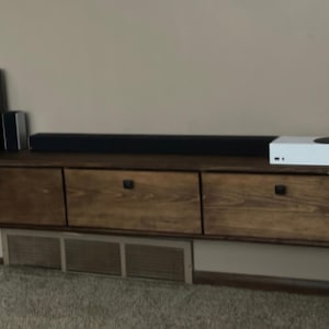 May include: A wall-mounted floating media console made of dark stained wood with three horizontal drawers, each featuring a small black handle. A black soundbar, a white gaming console, and two speakers are positioned on the top surface.