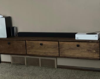 May include: A wall-mounted floating media console made of dark stained wood with three horizontal drawers, each featuring a small black handle. A black soundbar, a white gaming console, and two speakers are positioned on the top surface.