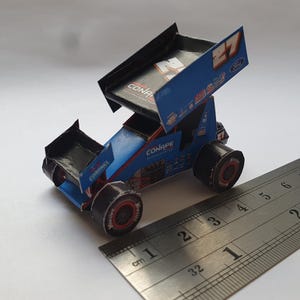 Può includere: Un modello di carta di un'auto da corsa blu e nera con il numero 27 sul lato. L'auto è appoggiata su un righello con segni in centimetri e pollici.