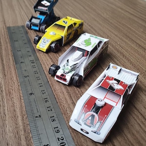 Puede incluir: Cuatro coches de carreras en miniatura hechos de papel. Los coches están pintados en diferentes colores, incluyendo amarillo, blanco, rojo y azul. Los coches tienen diferentes números, incluyendo 2, 23 y 7. Los coches están sentados sobre una superficie de madera.