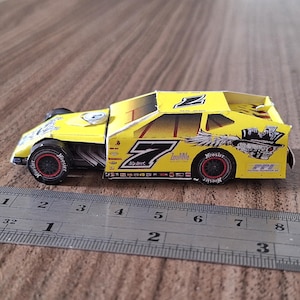 Pode incluir: Um modelo de papel amarelo de um carro de corrida com o número 7 no lado. O carro tem detalhes em preto e vermelho e as palavras "Monster" e "Zoom-Mile" no lado. O carro tem 17,8 cm de comprimento.