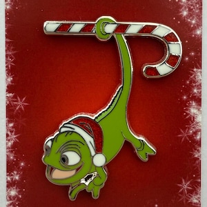 May include: A Christmas-themed enamel pin featuring a green chameleon wearing a Santa hat, holding a candy cane. The pin is set against a red background with the words "Merry Christmas" and snowflake accents.