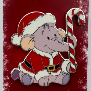 Puede incluir: Pin navideño con un elefante de dibujos animados vestido de Papá Noel, sosteniendo un bastón de caramelo. El fondo rojo tiene la inscripción "Merry Christmas" y detalles de copos de nieve.