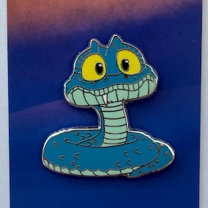 May include: A blue enamel pin featuring a cartoon snake with large yellow eyes, a wide grin, and a single visible white fang. The snake has a light green underbelly and is coiled. The text @elizapins is printed in the top left corner.