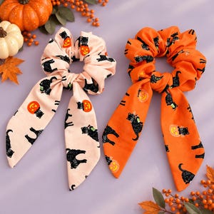 May include: Two fabric hair scrunchies with long ribbon tails. One scrunchie is light pink and the other is bright orange. Both feature a repeating pattern of black cats and orange jack-o'-lantern pumpkins printed across the fabric surface.