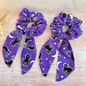 May include: Two purple fabric hair scrunchies featuring a repeating pattern of black cats, white crescent moons, small white stars, and black bats. Each scrunchie includes two long, dangling fabric tails attached to the elasticized ring.