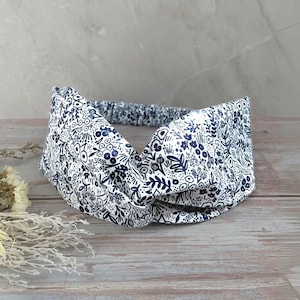 May include: A white headband with a navy blue floral pattern. The fabric is twisted at the front, and the back is an elastic band. The floral design includes various shapes of flowers and leaves.