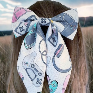 May include: A large hair bow featuring a white background with a pattern of video game controllers, headphones, and circuit board designs in gray, pink, and teal. The bow is tied in the hair.