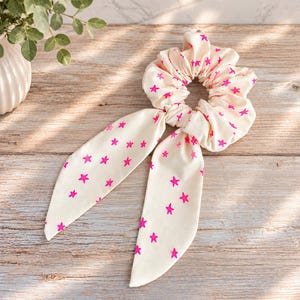 May include: A cream-colored fabric hair scrunchie featuring a repeating pattern of small, vibrant hot pink stars. The accessory includes two long, pointed fabric tails that hang down from the gathered elastic band, creating a decorative effect.