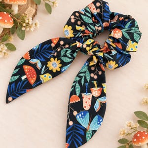 May include: A black fabric hair scrunchie featuring a colorful pattern of orange and white mushrooms, yellow flowers, blue butterflies, and green leaves. The scrunchie includes two long, pointed fabric tails that hang down from the gathered center.