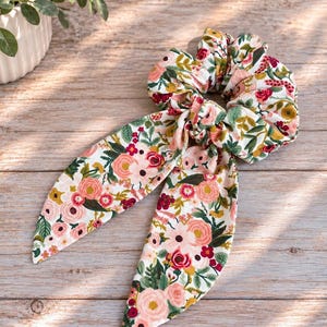 May include: A fabric hair scrunchie featuring a white background with a colorful floral pattern of pink, red, and yellow flowers along with green leaves. The scrunchie includes two long, pointed fabric tails that hang down from the center knot.