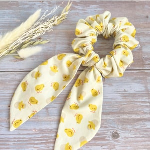 May include: A cream-colored scrunchie with a yellow chick pattern and long, matching fabric ties. The scrunchie is made of soft fabric and is suitable for various hairstyles. The design features small, yellow chicks on a cream background.