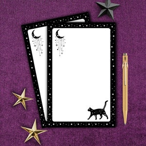 May include: Two black framed notepads with a white background and a black cat, crescent moon, and stars design. The notepads are on a purple background with gold and black stars.