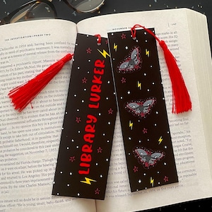 May include: Two black bookmarks with red tassels are placed in an open book. The bookmarks have a repeating star pattern, the text "LIBRARY LURKER", and moth illustrations. A pair of eyeglasses rests on the book.