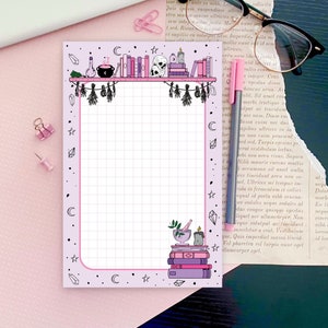 May include: A pink and white gridded notepad with a black and white illustration of a bookshelf with books, candles, a cauldron, and skulls. The border of the notepad is decorated with stars, moons, and crystals.