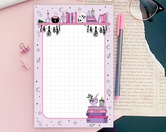 May include: A pink and white gridded notepad with a black and white illustration of a bookshelf with books, a cauldron, a skull, and candles. The illustration is surrounded by a border of stars, moons, and crystals. The notepad has a pink background and a white grid.