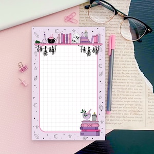 May include: A pink and white gridded notepad with a black and white illustration of a bookshelf with books, a cauldron, a skull, and candles. The illustration is surrounded by a border of stars, moons, and crystals. The notepad has a pink background and a white grid.