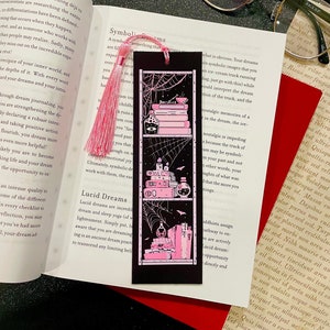 May include: A black bookmark featuring a pink and white illustration of a bookshelf with books, candles, and potion bottles. The bookshelf is decorated with a spiderweb and bats. The bookmark has a pink tassel.
