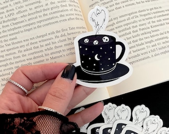 May include: Black and white sticker featuring a spooky coffee cup design. The cup is adorned with skulls, stars, and a crescent moon, with a ghost rising from the top. A stack of similar stickers is visible.