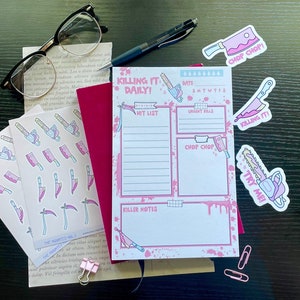 May include: A pink and white planner with the title "Killing It Daily!"  The planner has sections for a hit list, urgent kills, and killer notes. There are also several pink and white stickers with images of chainsaws and knives.