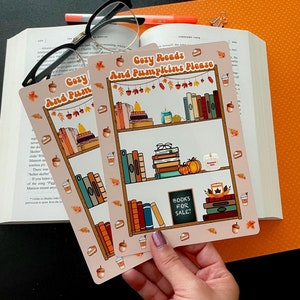 May include: Two pink and white printable stickers with a fall theme. The stickers feature a bookshelf with books, pumpkins, and candles. The text on the stickers reads "Cozy Reads and Pumpkins Please" and "Books for Sale!"