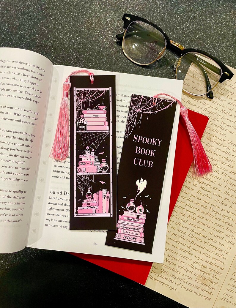 May include: Two black bookmarks with pink and white illustrations of books, potions, and a ghost. The bookmarks have pink tassels and the text "Spooky Book Club" on one bookmark and "Witch Craft, Black Magic, Poisons" on the other.